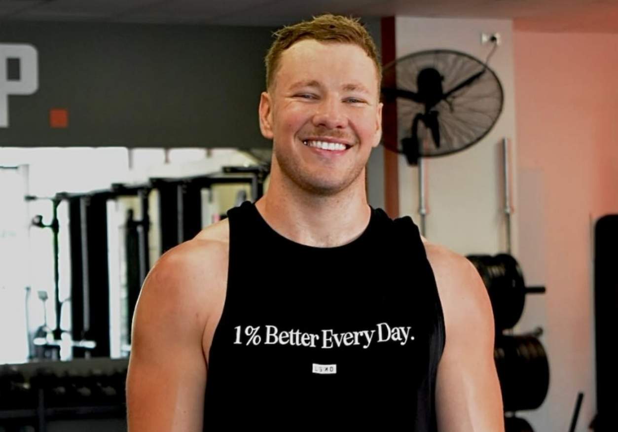 Josh, who lives with hypertrophic cardiomyopathy, is smiling at the gym, with a tank top with slogan '1% Better Every Day'