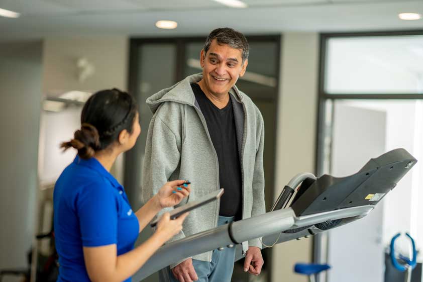 Older male in exercise gear smiling, speaking with a physiotherapist on a walking machine