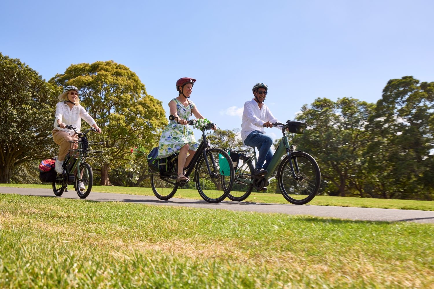 People wearing helmets ride bicycles along a sunny park path, smiling as they cycle together outdoors.