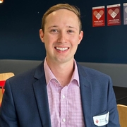 Dr. Lachlan Dalli, in a suit and checkered shirt, sits smiling in a modern office space