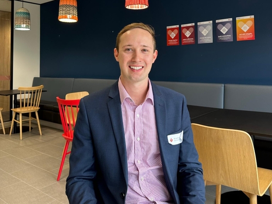 Dr. Lachlan Dalli, in a suit and checkered shirt, sits smiling in a modern office space