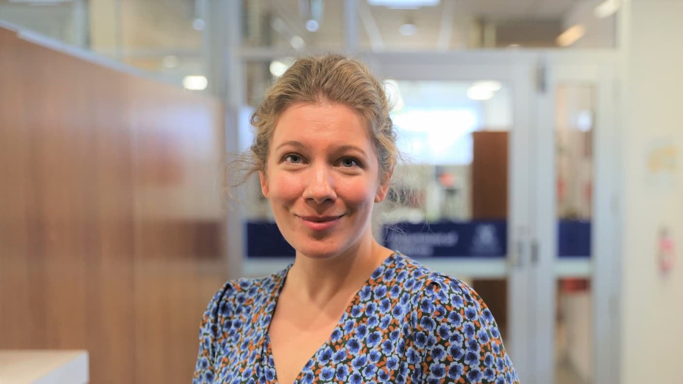 Dr Elizabeth Paratz, stands indoors in a blue patterned dress, smiling with glass doors and office furniture visible in the background at St Vincent's Institute of Medical Research.