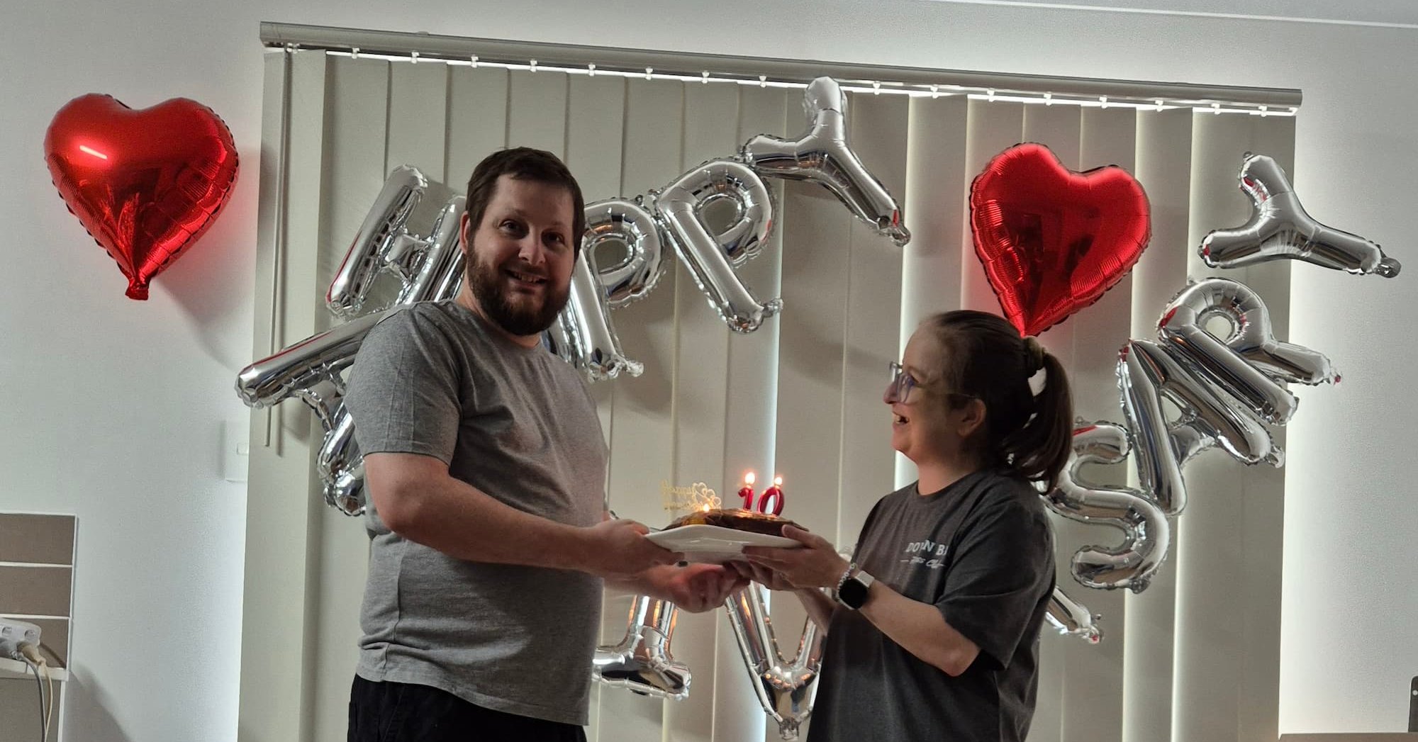 Two people holding a birthday cake indoors, with heart‑shaped balloons and a “Happy” balloon display in the background