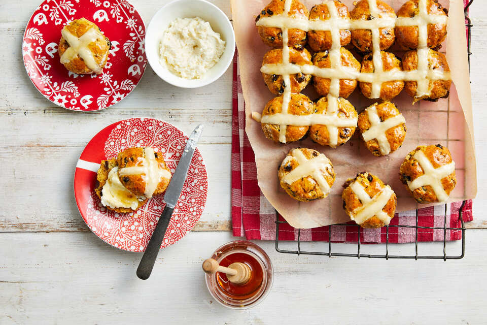 White table set with plates, butter, jam dishes and a serve of 24 hot cross buns resting on the red and white tea towel 