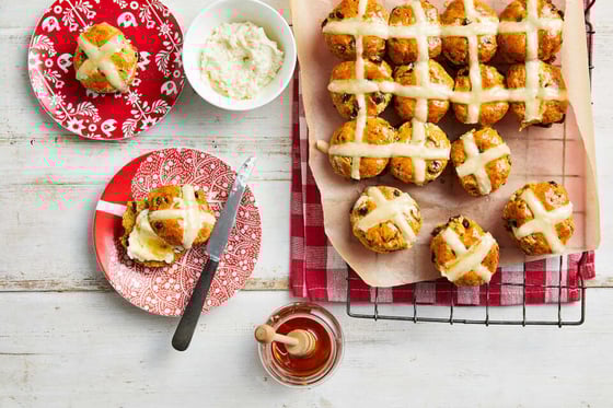 White table set with plates, butter, jam dishes and a serve of 24 hot cross buns resting on the red and white tea towel