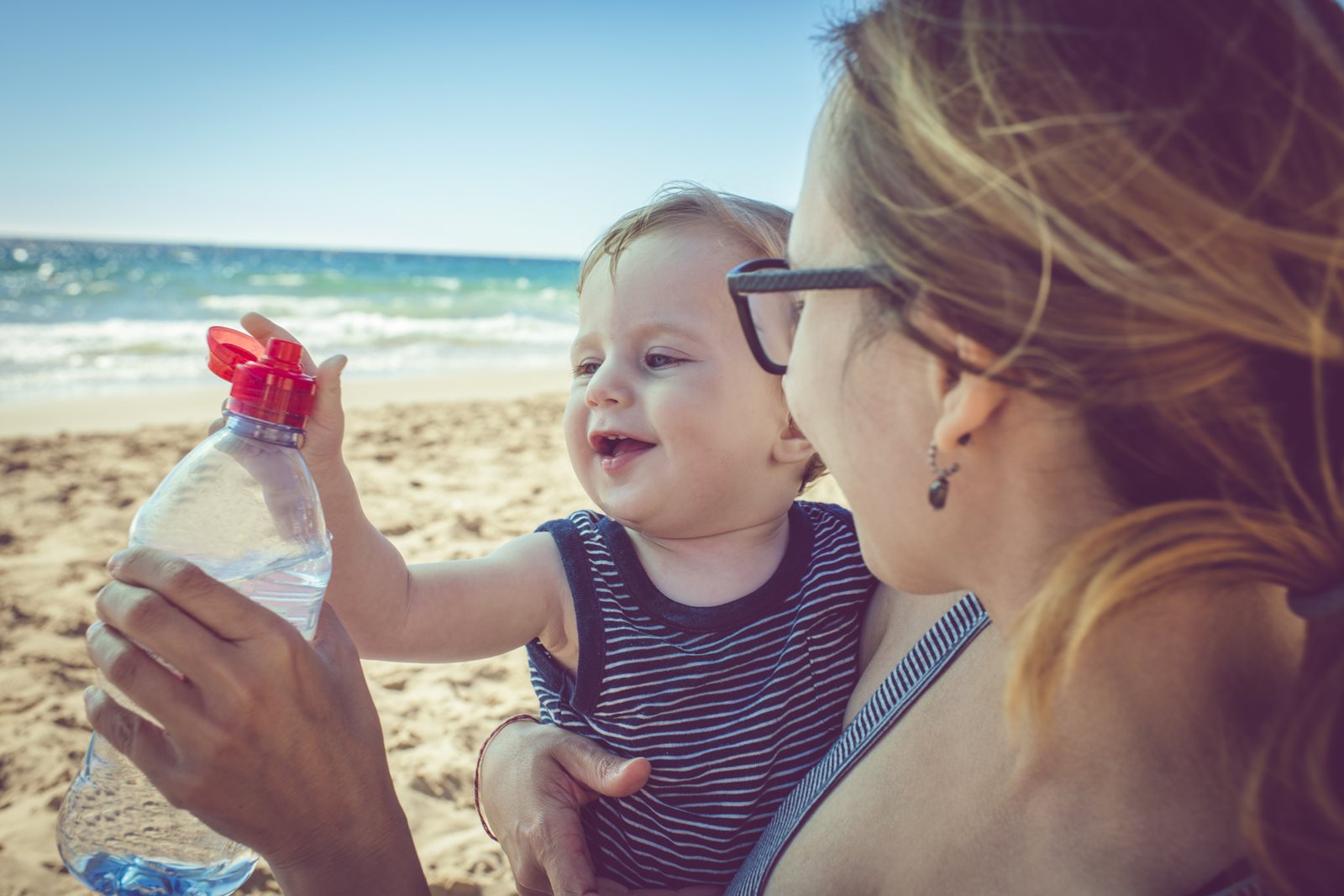 A woman cradling a baby on a beach, offering a bottle of water.