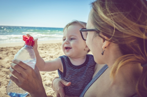 A woman cradling a baby on a beach, offering a bottle of water.