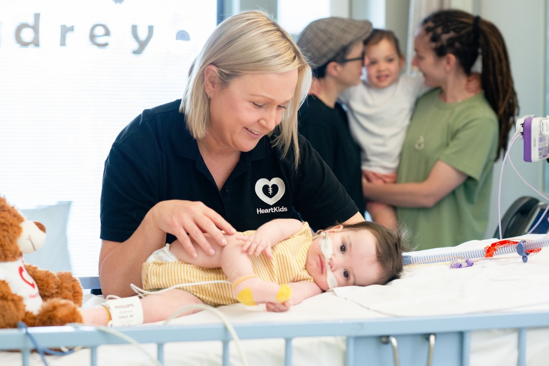 A HeartKids worker supporting a young infant who is in hospital with childhood onset heart disease. The child's family are chatting and hugging in the background. 