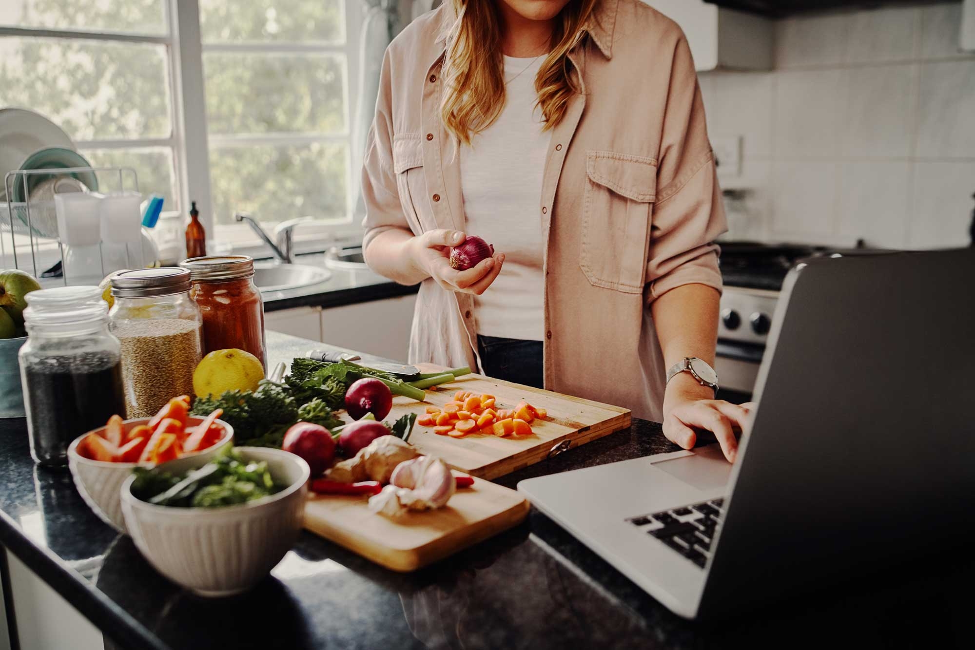 A woman is prepping food at her kitchen bench while looking at a recipe on her laptop
