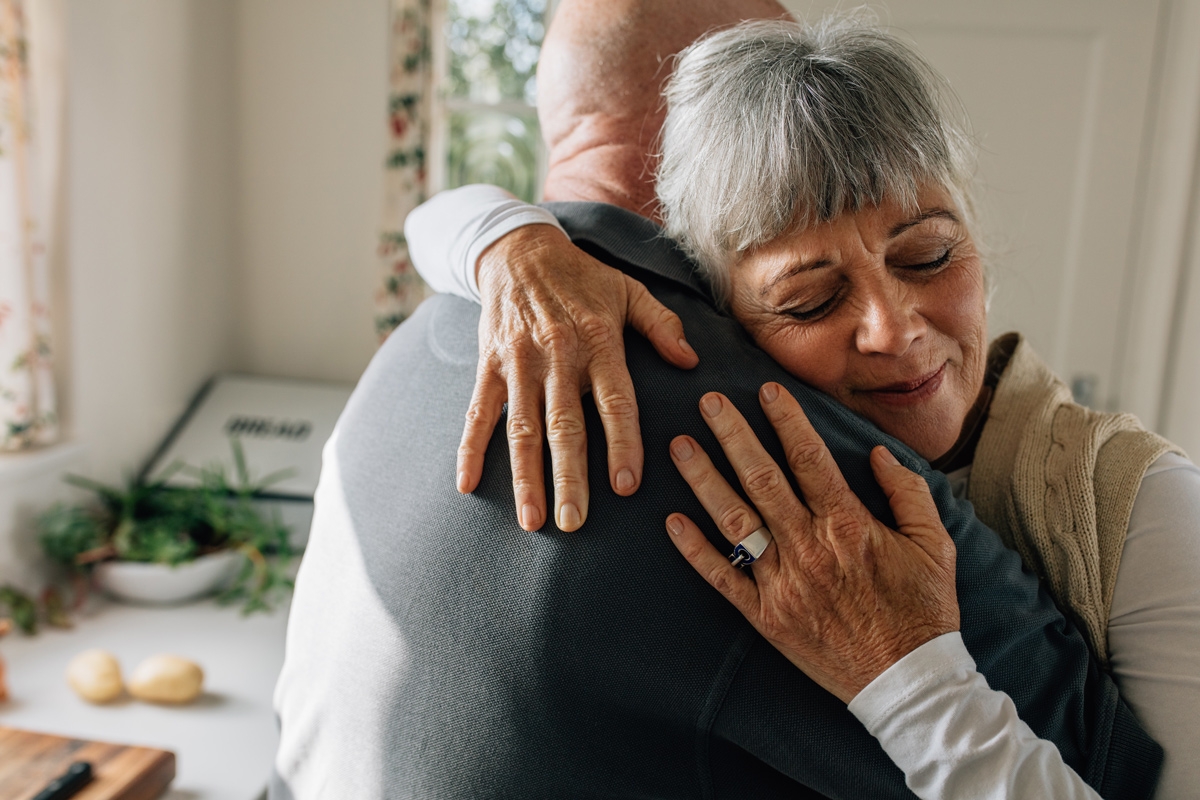 An elderly woman embraces her husband lovingly in their kitchen.