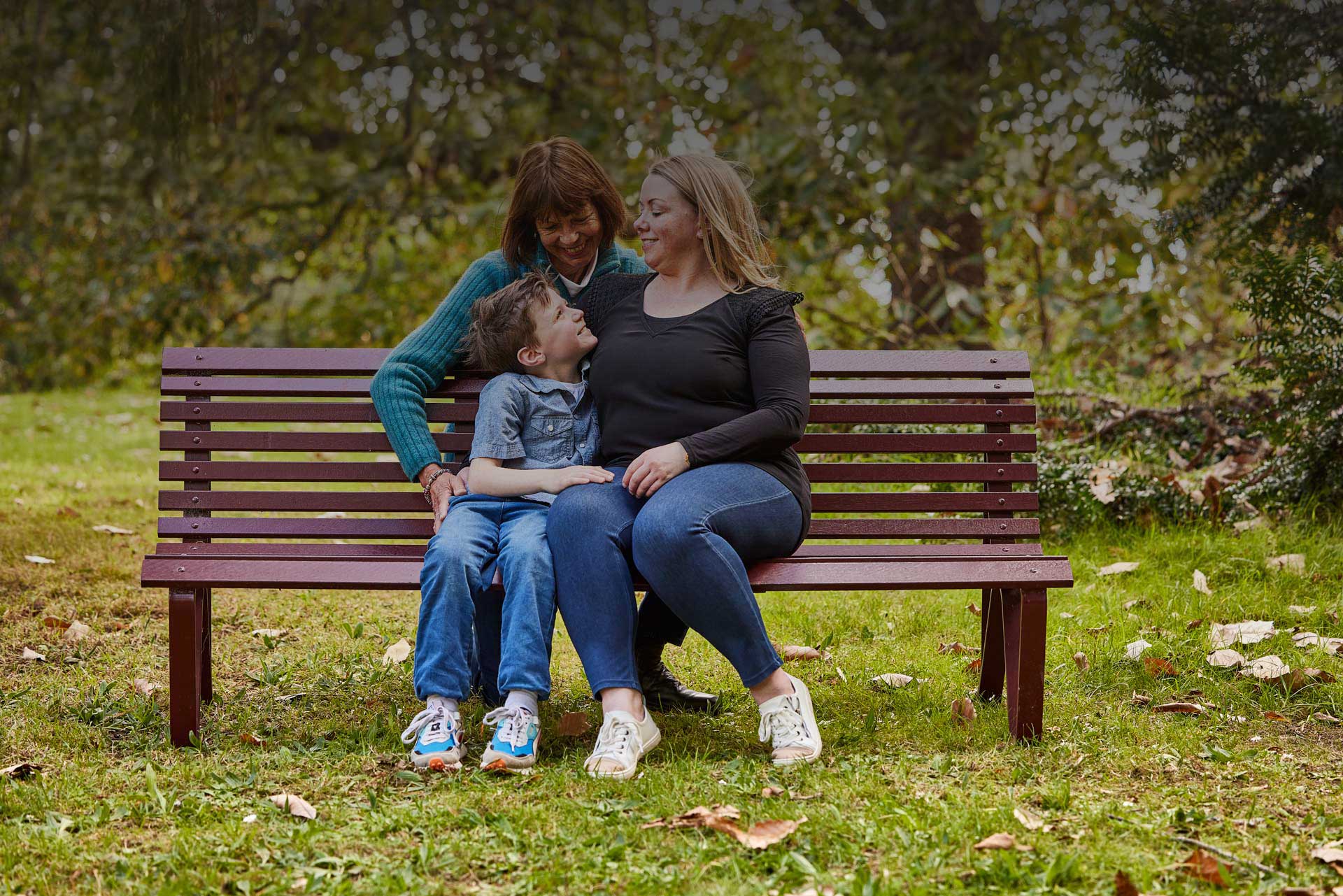 Family of three, older mum, daughter and son/grandson, sitting outdoors in a leafy park on park bench