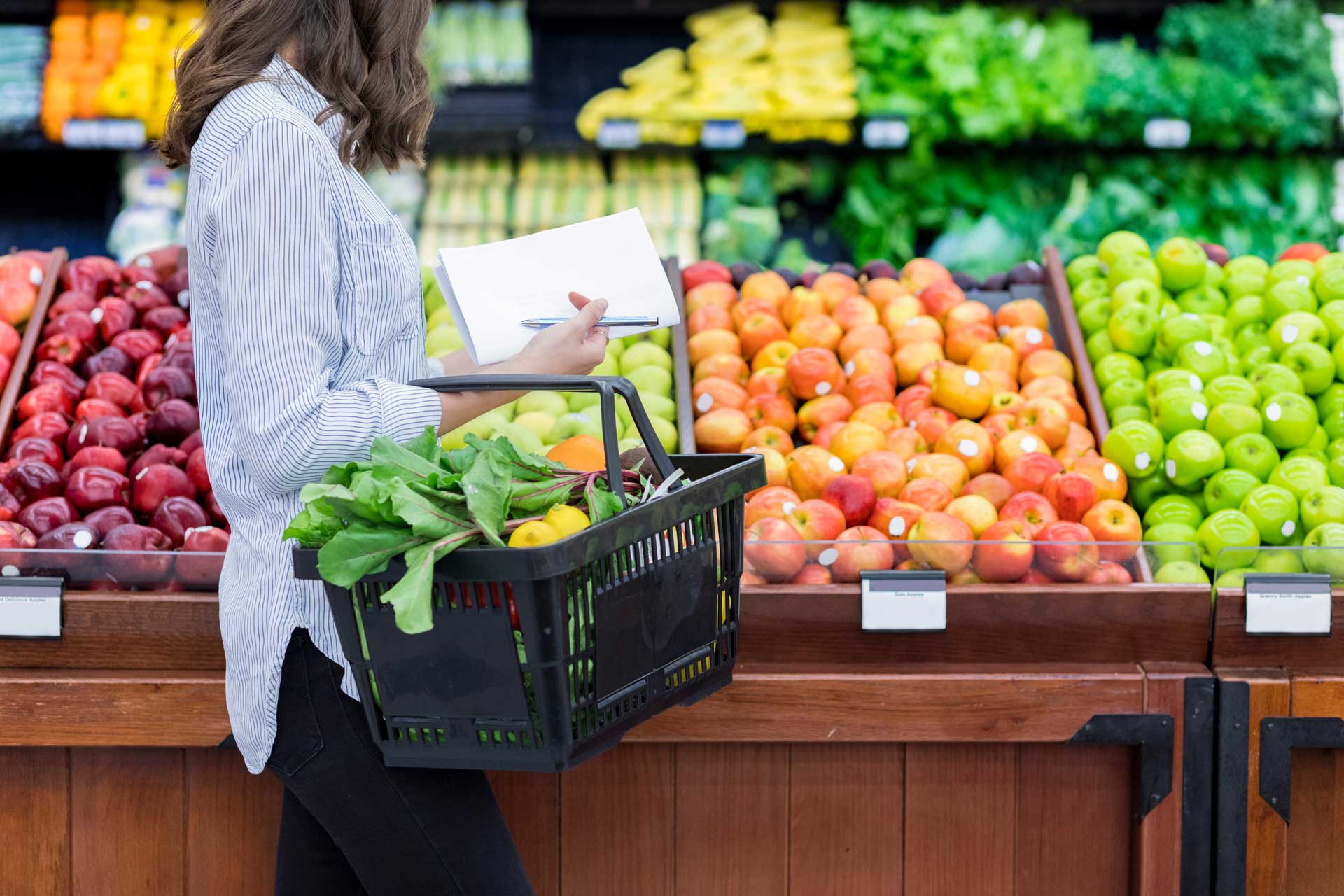 Woman in fruit and veg section of grocery store, holding a shopping list and basket 