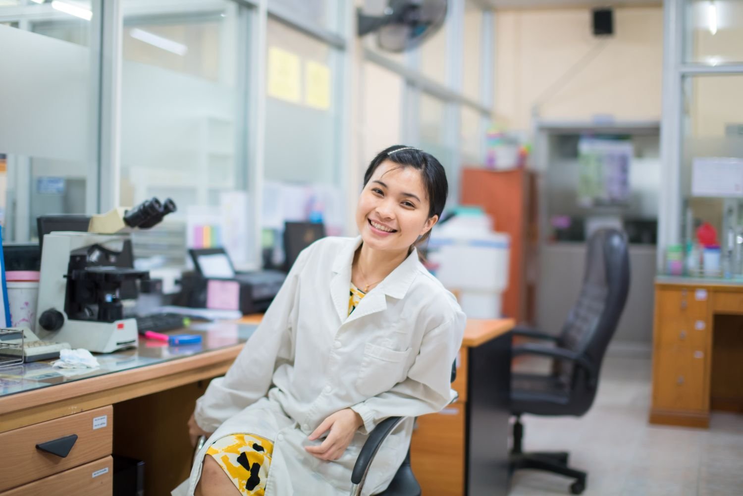 Smiling researcher in a white lab coat seated beside a microscope in a laboratory workspace.