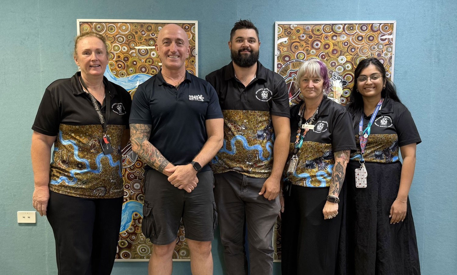 Five people standing together indoors in front of colourful Aboriginal artworks, with four wearing matching patterned shirts.