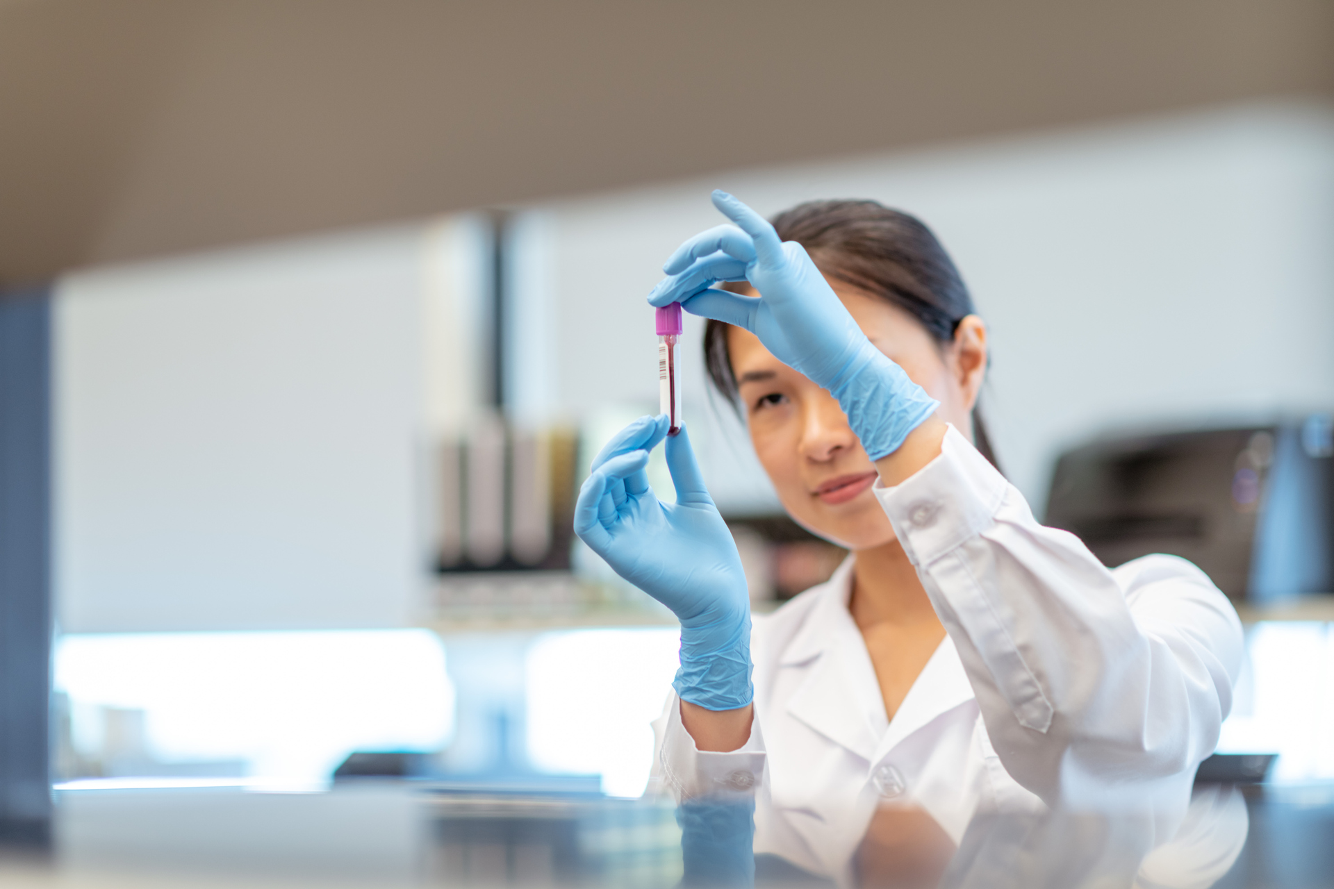 A scientist in a lab coat and blue gloves examines a blood sample in a test tube for a blood test.