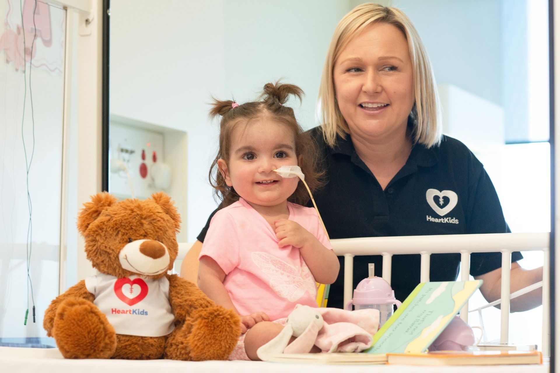 A HeartKids volunteer standing next to the hospital bed of a young girl, both are smiling at the camera