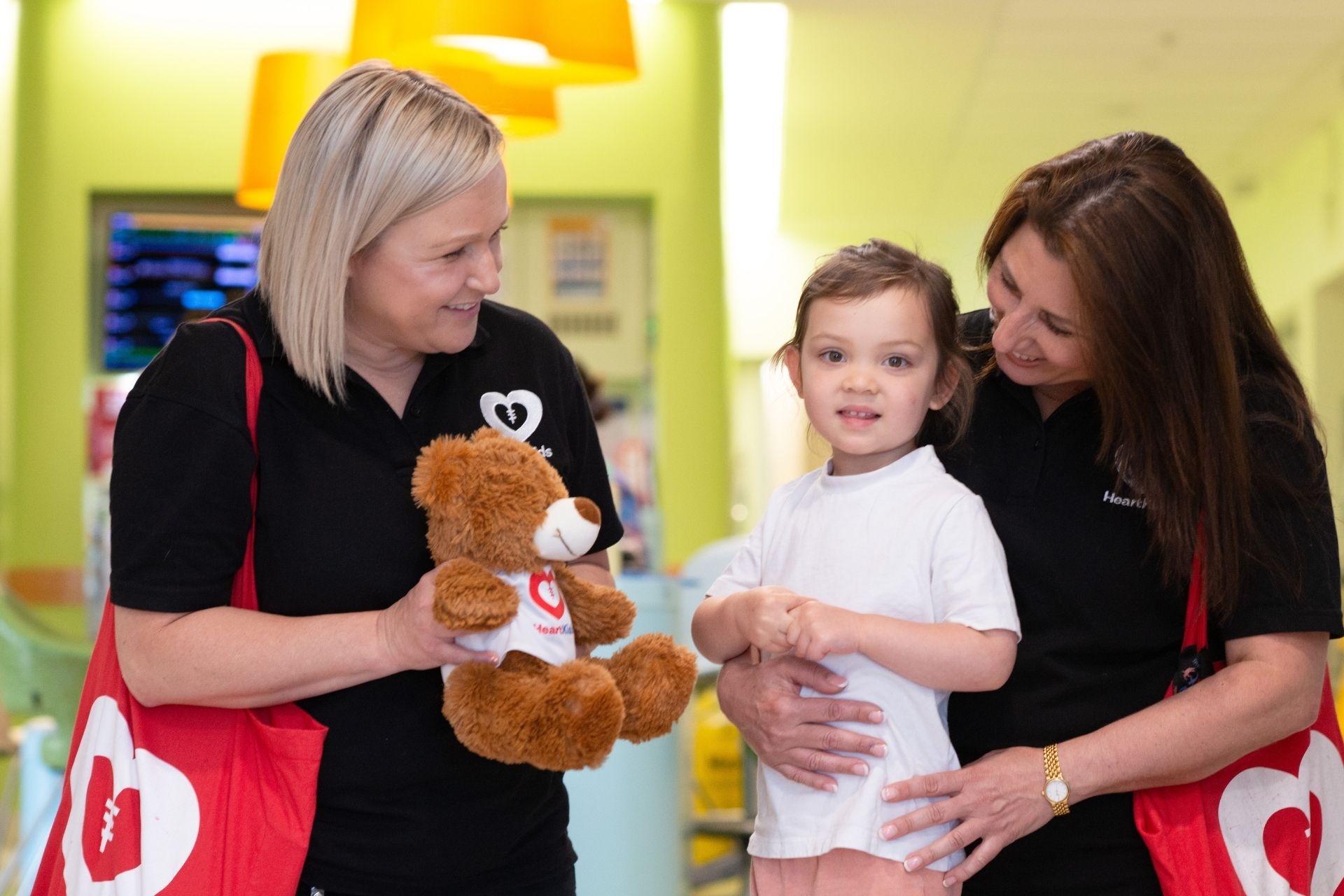 A HeartKids staff member handing a teddy bear to a small girl at the hospital