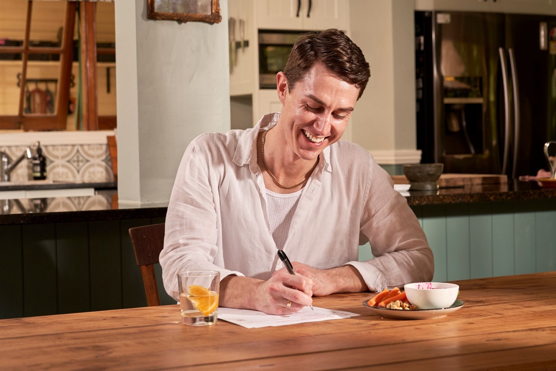 A young man writing a shopping list at a kitchen table. He's eating a plate of healthy snacks and has a glass of water with an orange in it. 