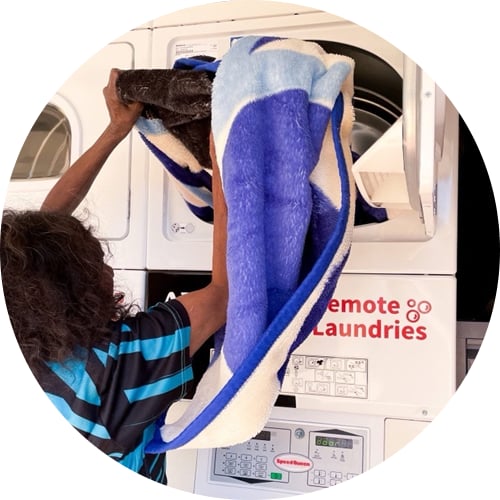 Person placing a large blue and white blanket into a commercial dryer at a remote laundry facility.