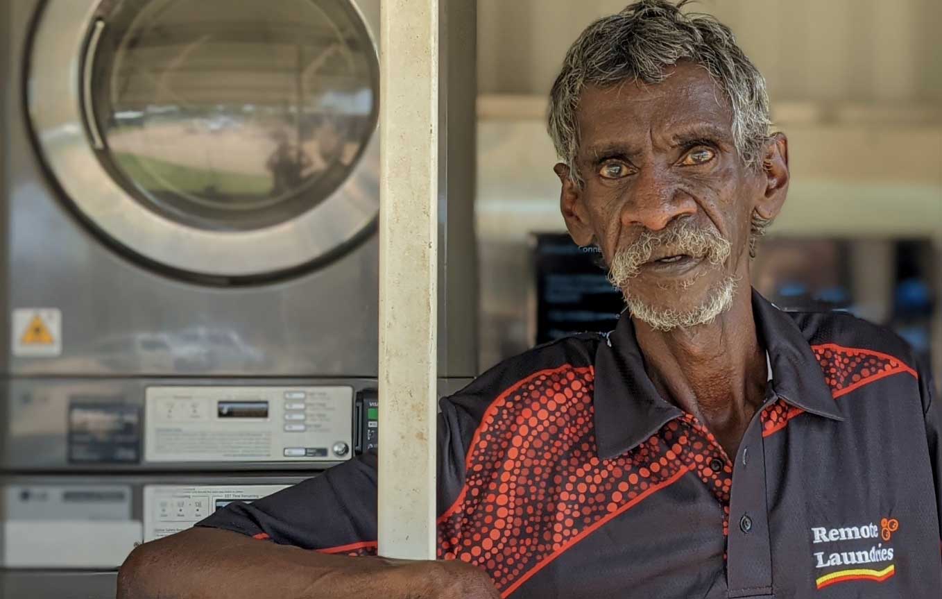 A First Nations man wearing a red shirt is positioned in front of a washing machine.