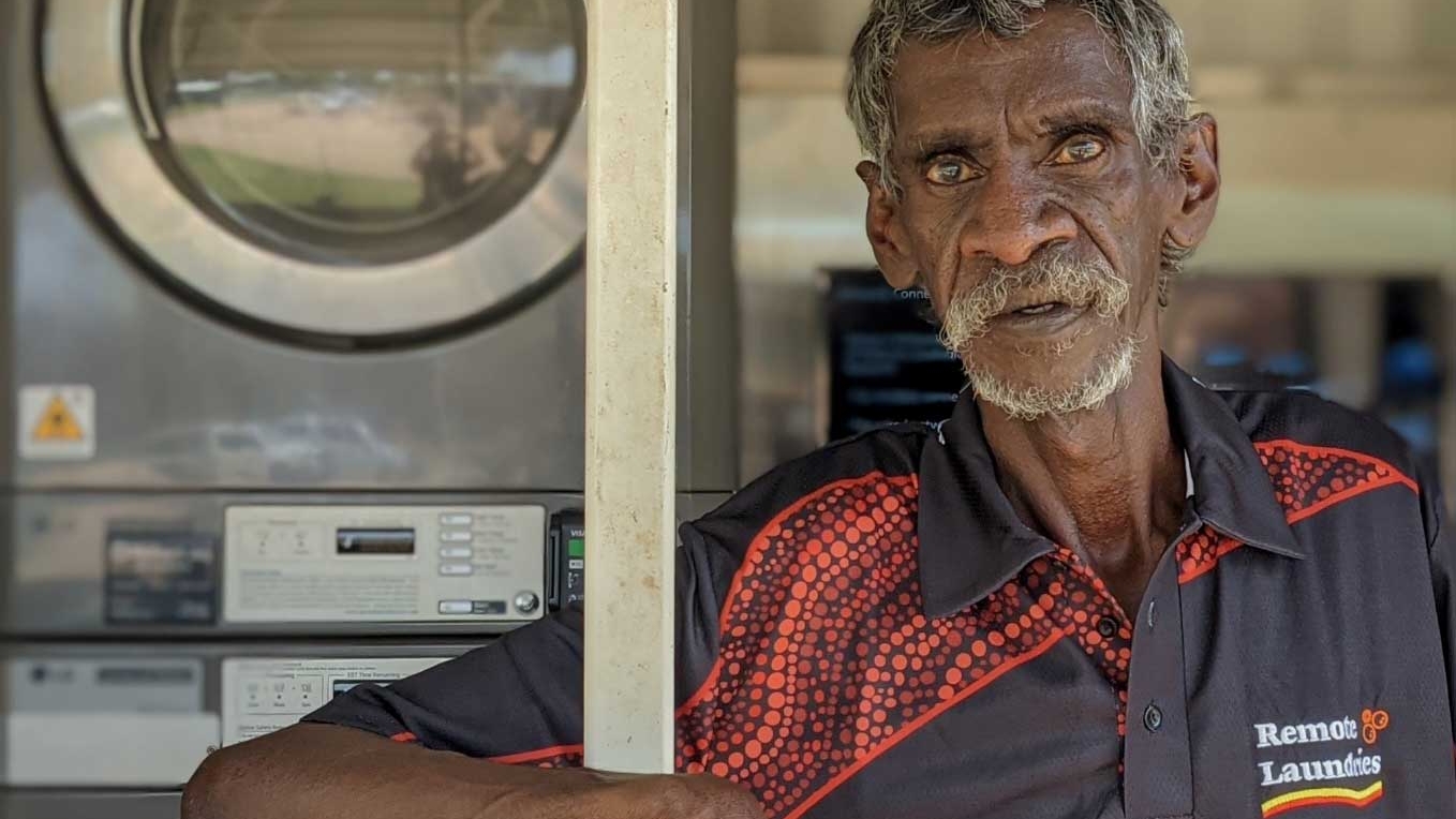 A First Nations man wearing a red shirt is positioned in front of a washing machine.