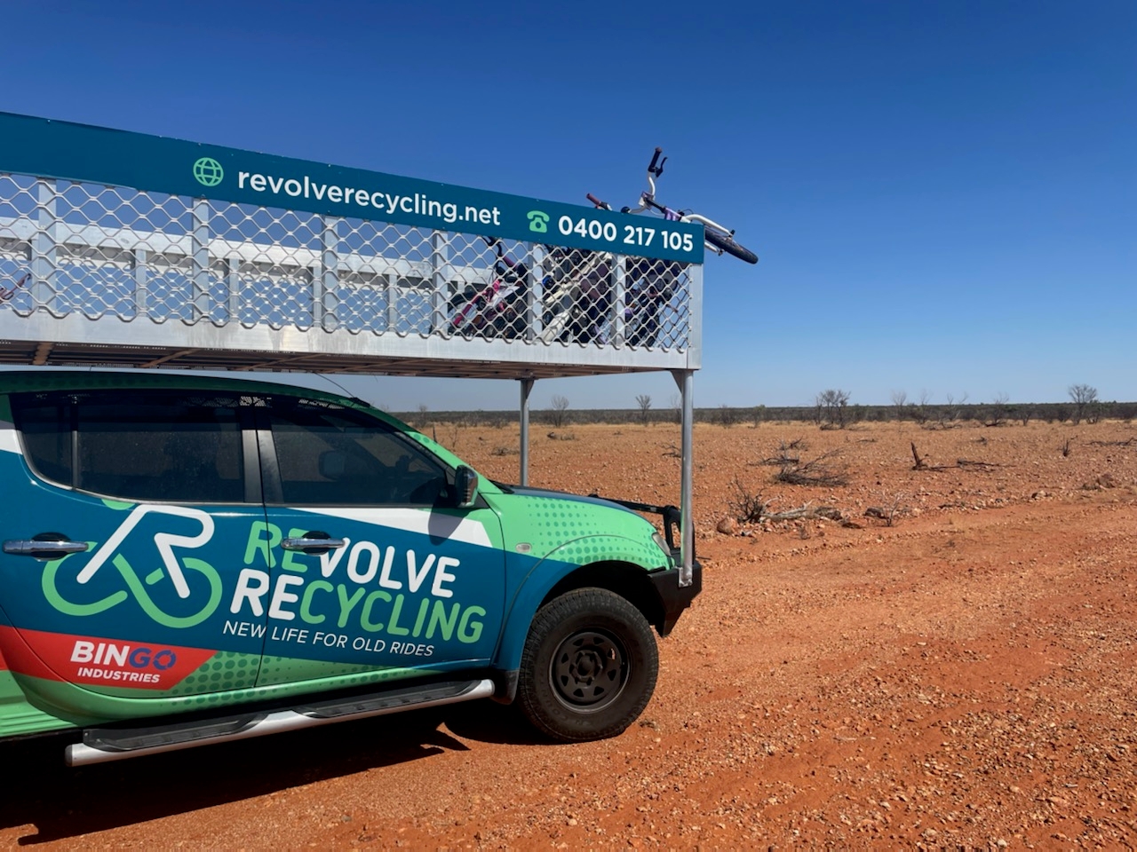 A blue and green truck with 'Revolve Recycling' written on the side parked in the Australian desert. The truck is carrying an overhead tray with bicycles in it, for communities to use.  