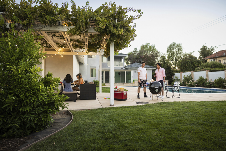 A modern backyard featuring a lush green lawn, a swimming pool, and a pergola covered in climbing vines and string lights. Two men stand by a charcoal grill while two women sit at a patio table, illustrating a functional outdoor entertainment area.