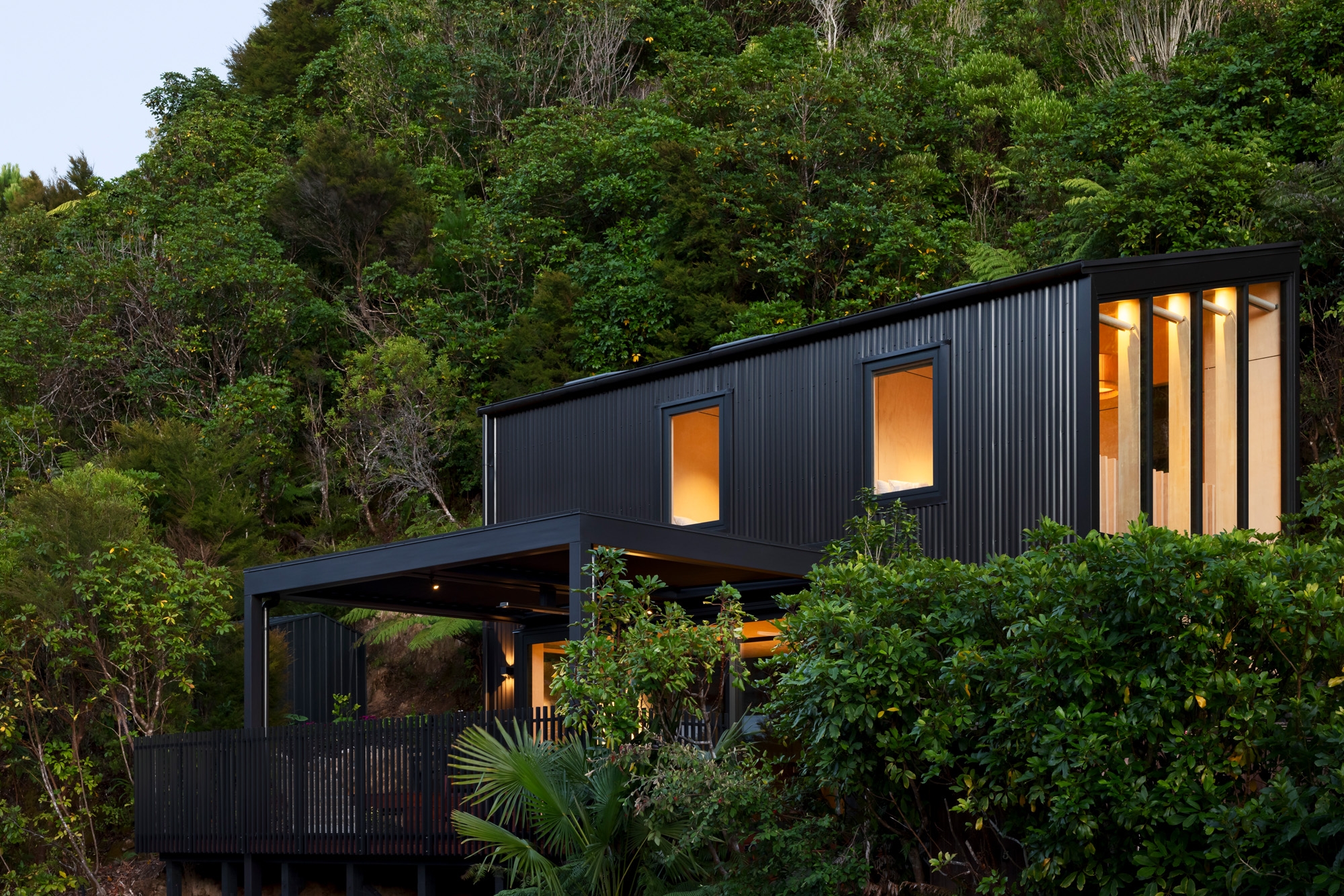 This image depicts a modern, two-storey house nestled within lush green foliage on a hillside. The house has a sleek, black exterior with a corrugated metal finish. The design features large windows that allow warm light to illuminate the interior, creating a cosy contrast against the dark facade. The house includes a spacious, elevated outdoor deck surrounded by a black railing, offering an open, sheltered area for relaxation. The dense greenery and trees surrounding the house give it a secluded, tranquil atmosphere, blending contemporary architecture with natural beauty.