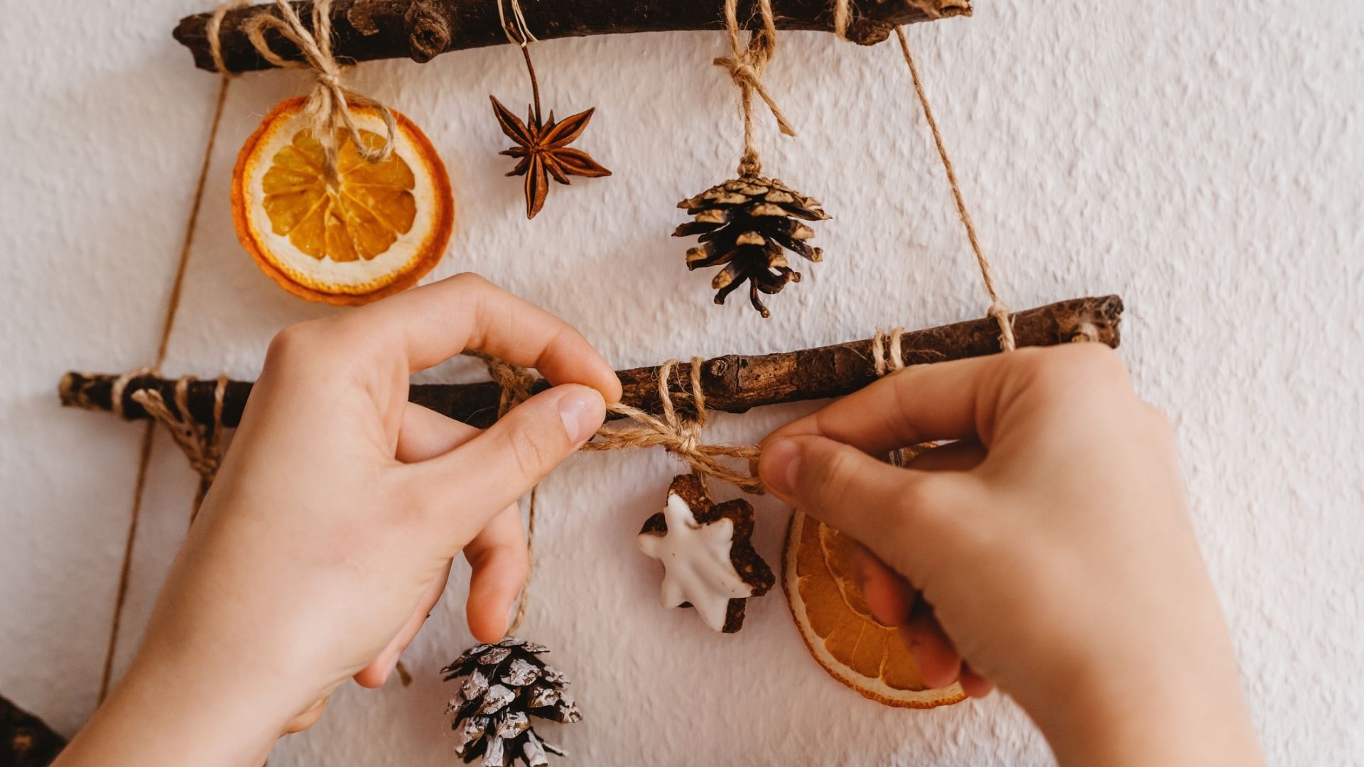 Two sticks in the shape of a Christmas tree hang on a wall, attached with twine are dried orange slices and pinecones. A pair of hands tie a bow on a piece of twine attaching a small cookie to one of the sticks.