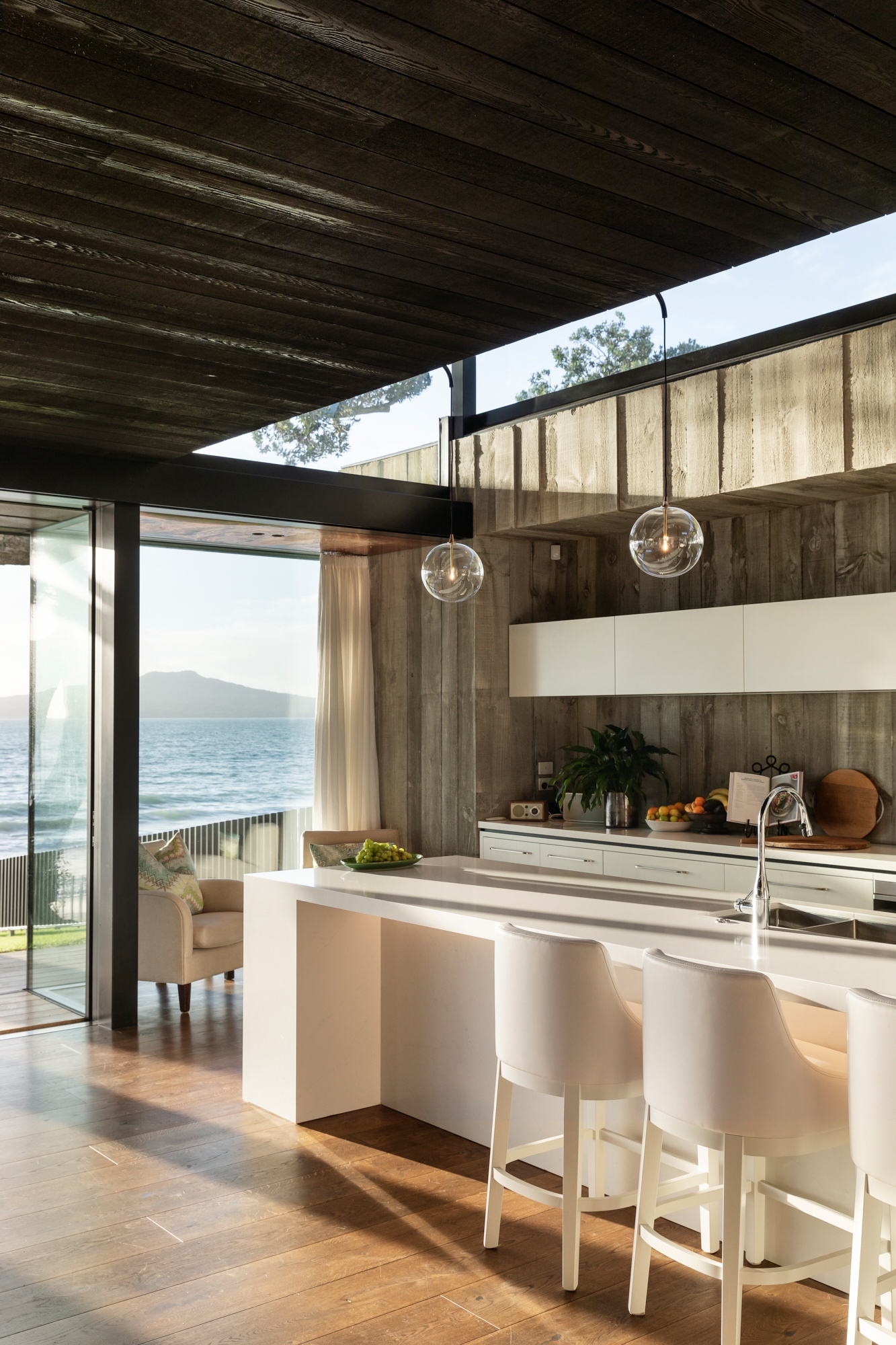 The image shows a modern kitchen with a sleek and minimalist design. The focal point is a large white island with a built-in sink and a high-gloss finish. Two contemporary white bar stools with curved backs are positioned at the island. The kitchen features a mix of natural and industrial elements, including wooden floors and walls with a raw, unfinished look. Large glass windows and sliding doors offer an unobstructed view of the ocean, filling the space with natural light. The ceiling has dark wooden panels, and two spherical pendant lights hang above the island, adding a touch of elegance. The background includes a cozy seating area with a cushioned chair and a small table, making the space inviting and comfortable.