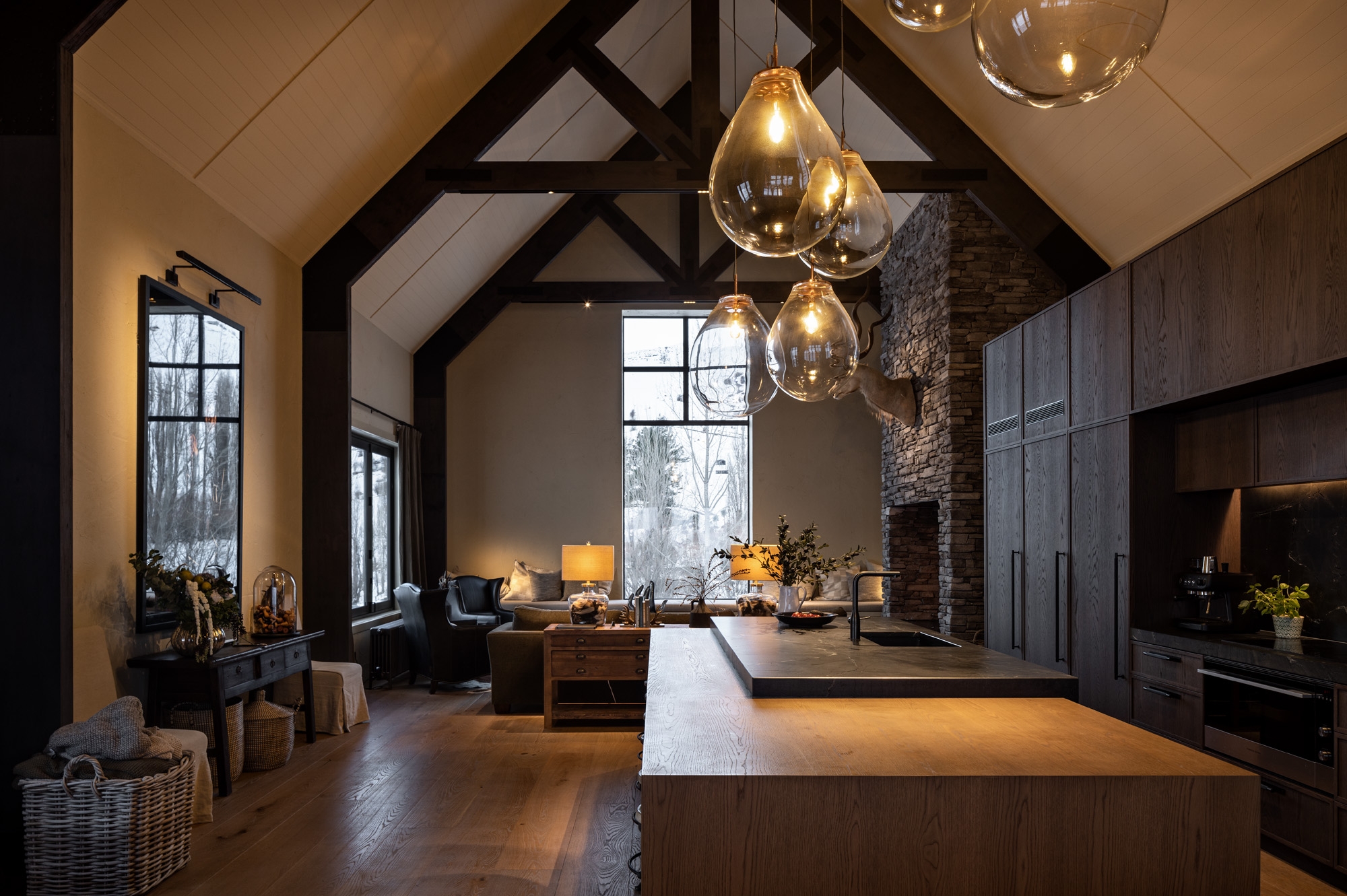 A modern rustic barn house kitchen with high ceilings, exposed wooden beams, dark cabinets, and pendant lighting. Large windows bring in natural light. A living area with a fireplace is visible in the background.