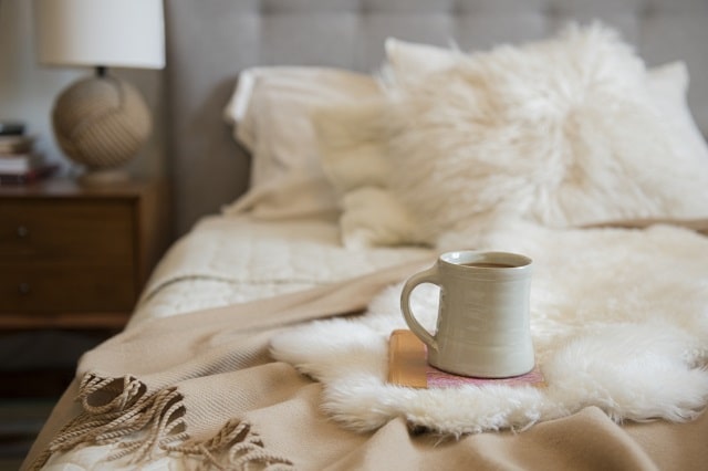  Ceramic mug of tea sitting on top of a book on a bed covered by different types of throws.