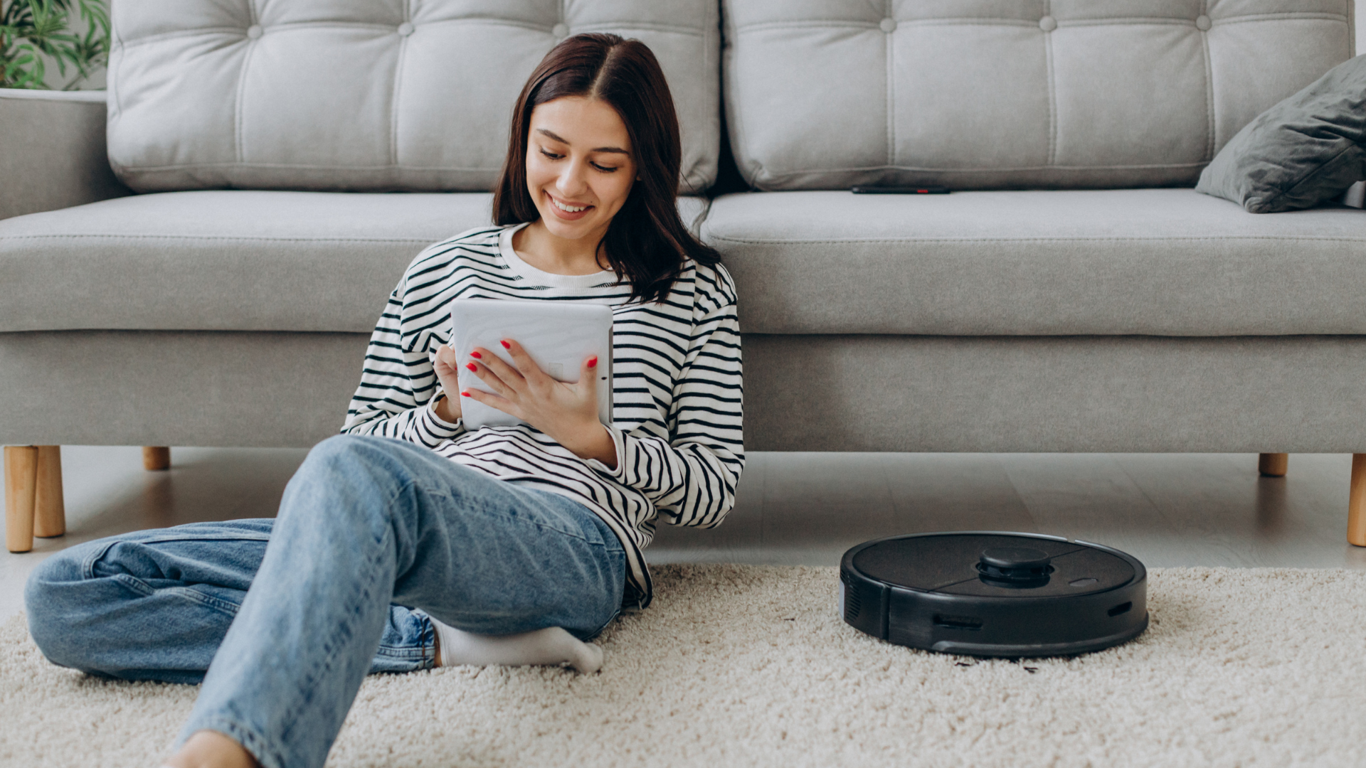 Woman sitting on ground leaning on couch next to robot vacuum cleaner