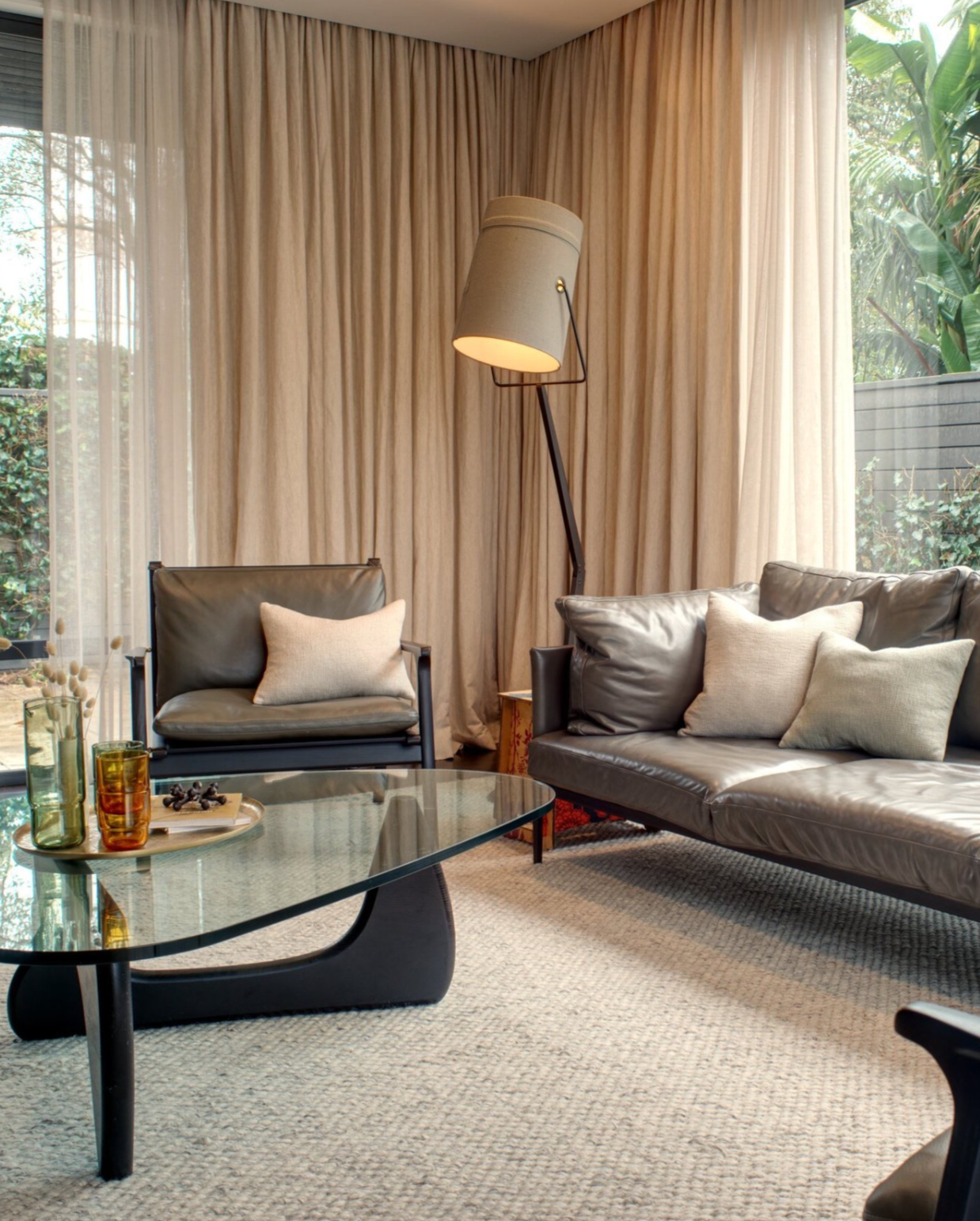Living room with layered, neutral curtains, leather armchairs, and a glass coffee table on a textured rug.