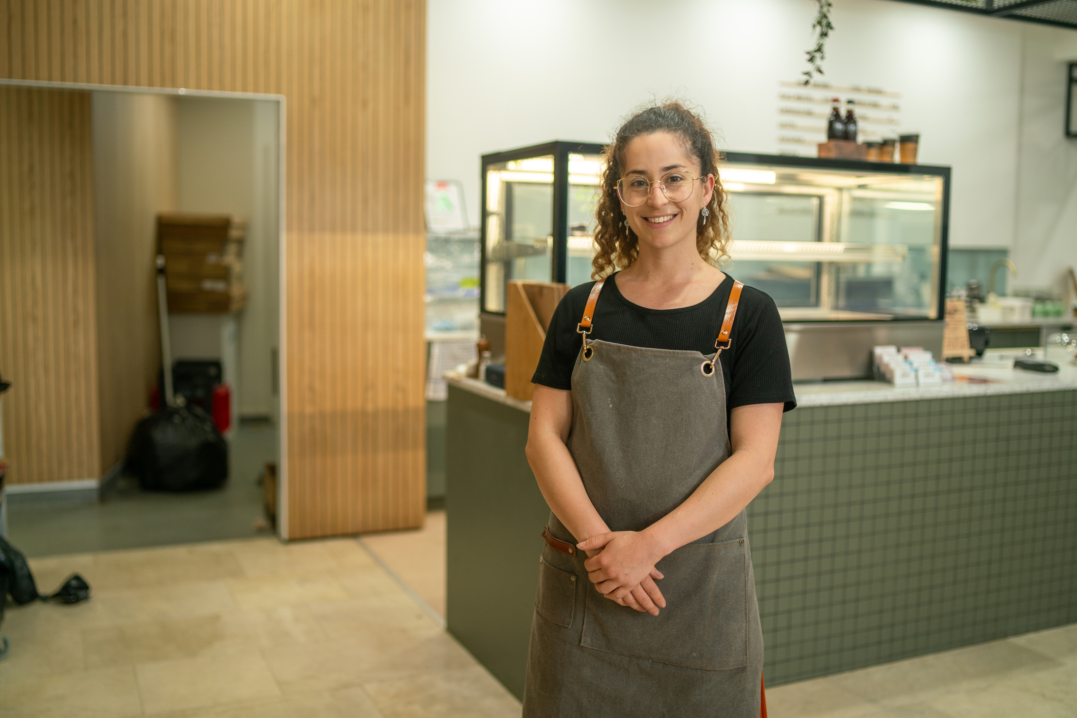 Young woman working in New Zealand counter, standing in front of cafe counter and cabinet.