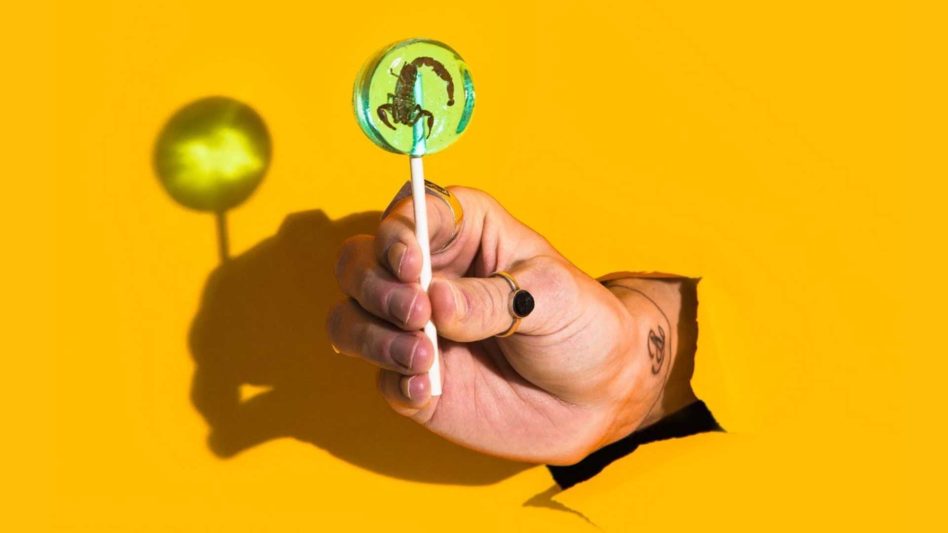 A hand pops out of a bright yellow background holding a lollipop with a scorpion inside