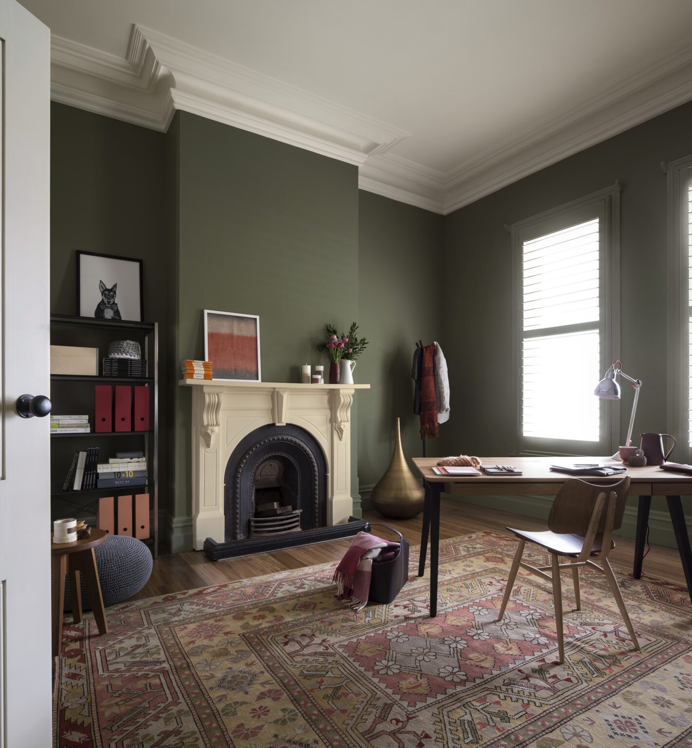 Dark green paint adorns the walls in this office. In the centre of the wall is a large white vintage fireplace complemented by a red Persian rug. 