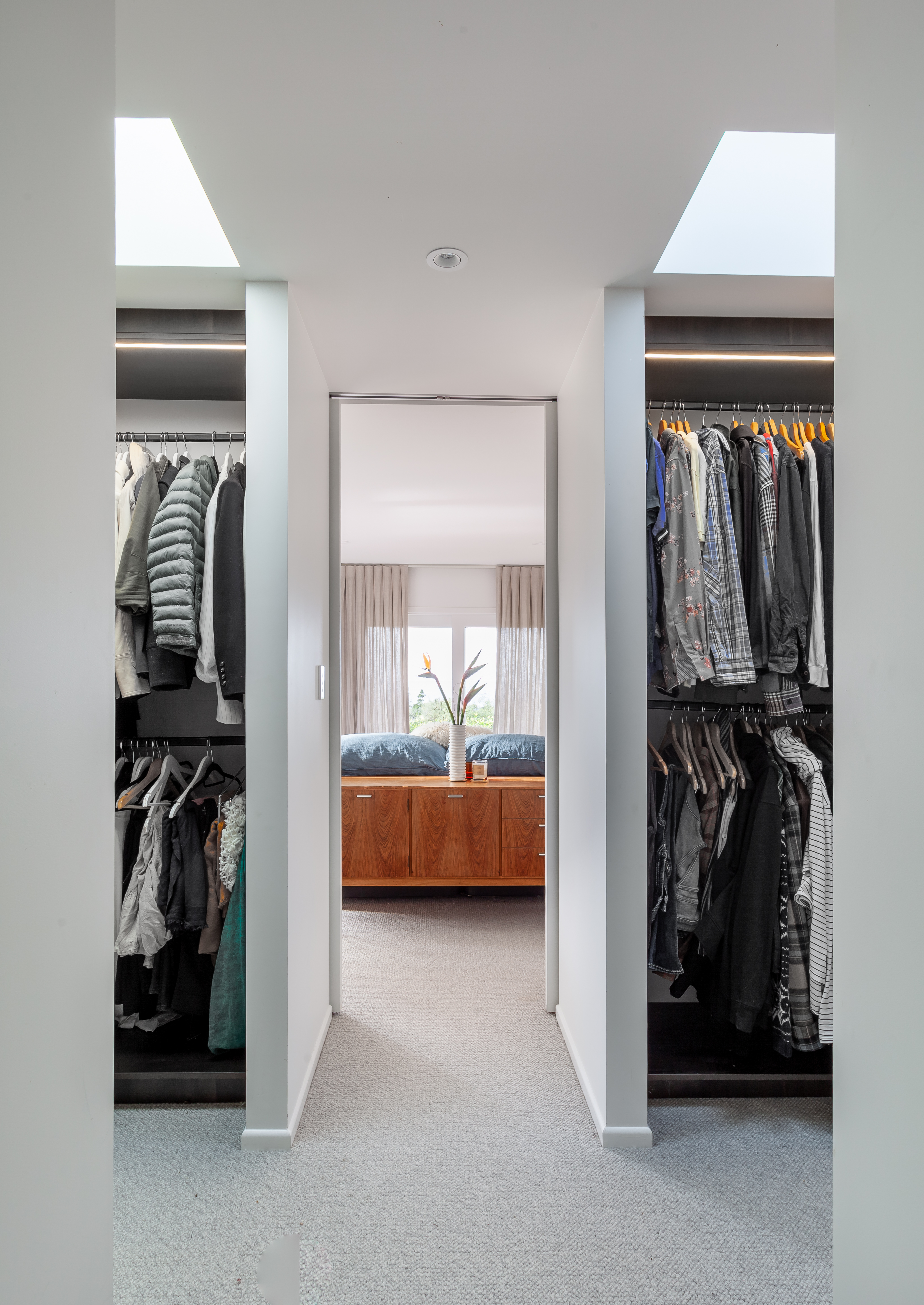 The image shows a modern, organized walk-in closet leading into a bright bedroom. On the left side of the closet, there are multiple grey-toned clothing items including shirts and jackets hung neatly. On the right side, various patterned and colored clothing items are displayed. The walls of the closet are white, creating a clean and minimalist look. The floor is covered with a light grey carpet that extends from the closet into the bedroom. In the background through an open doorway, part of a bedroom is visible featuring a large bed with two pillows and wooden detailing at its base. A window in the bedroom is partially visible; it is covered by light curtains allowing natural light to illuminate both rooms.