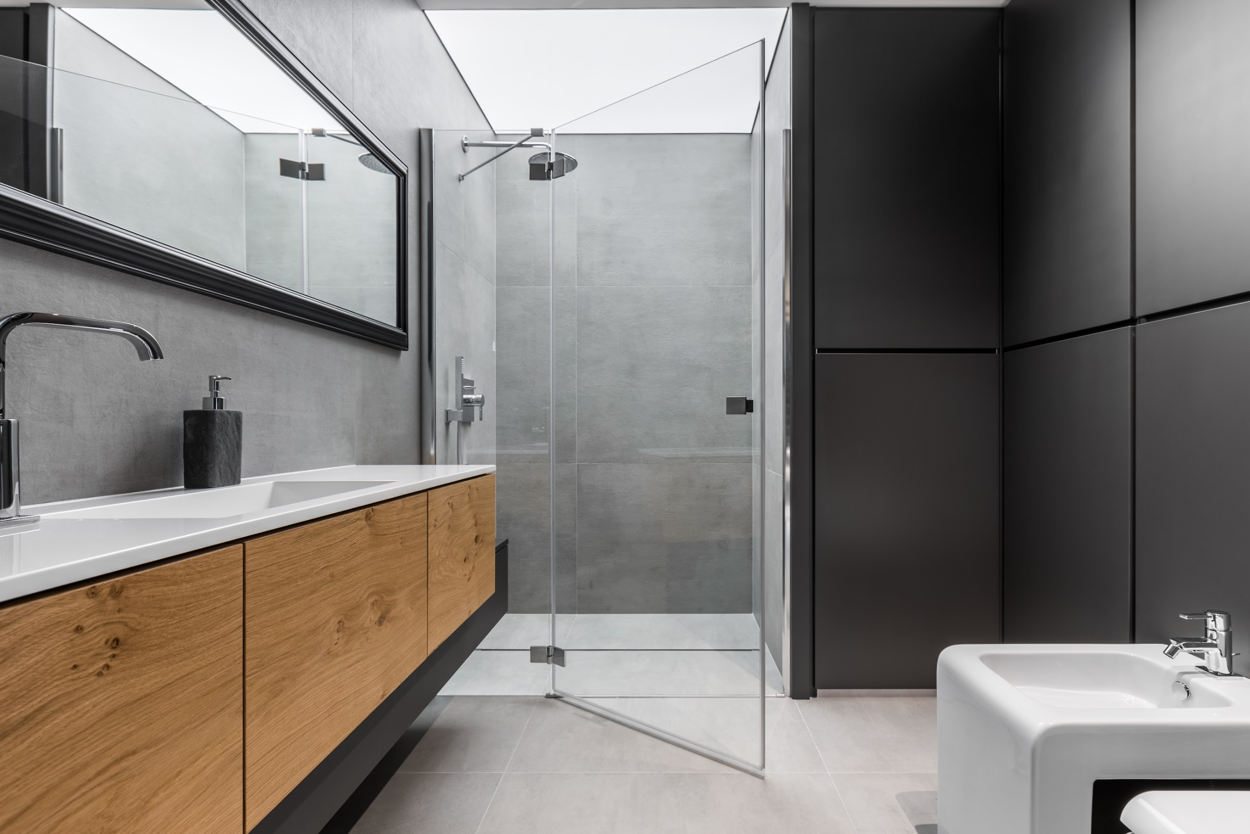 Modern bathroom with a minimalist design, featuring a large floating vanity with natural wood drawers, a rectangular mirror, and a spacious glass-enclosed shower. The room is accented with matte black cabinetry and light grey tiles.