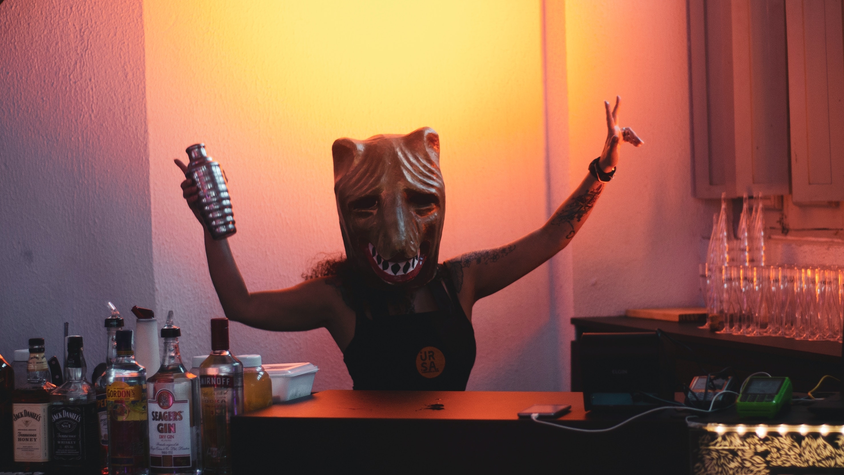 Woman in a scary animal mask dancing behind a bar holding up a cocktail shaker. 