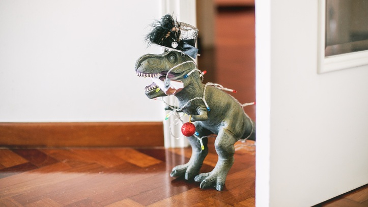 Dinosaur figurine standing on a wooden flor between a door and the wall. The dinosaur is decorated in Christmas lights.