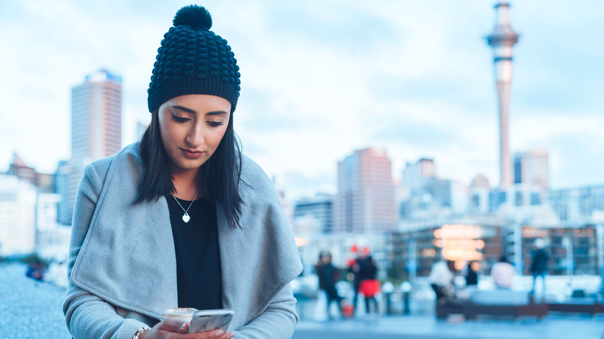 Young woman checks her phone to learn about the changes to the OCR in May 2024, in Auckland New Zealand. The Sky Tower is visible in the background.