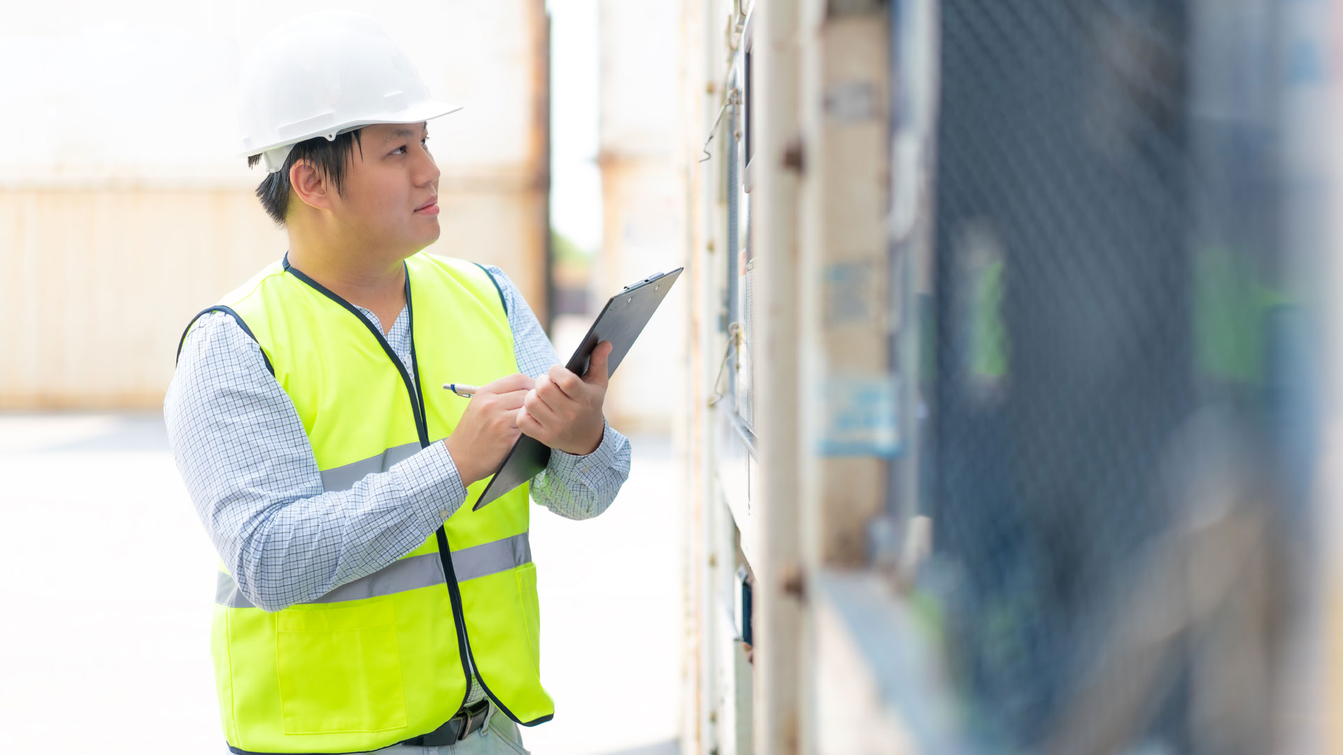 Foreman in a high vis and hard hat inspecting goods with a clipboard.