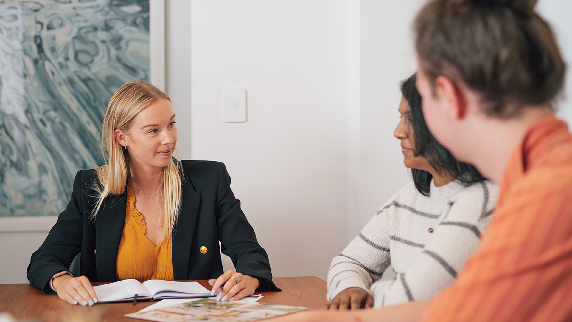 Real estate agent talking to a couple, sitting at a table with lots of brochures on it.