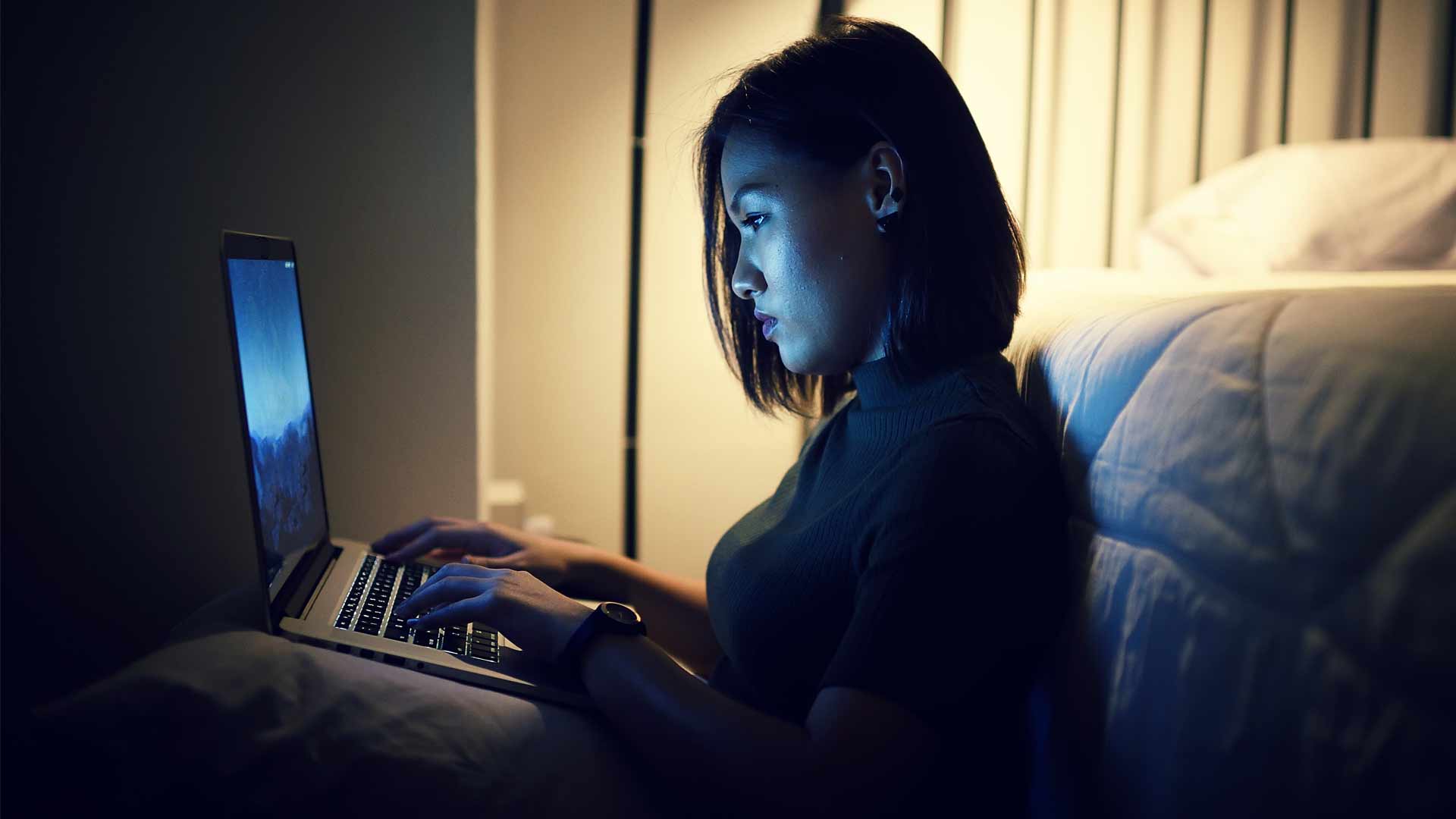 Woman sitting against her bed typing on her laptop late at night