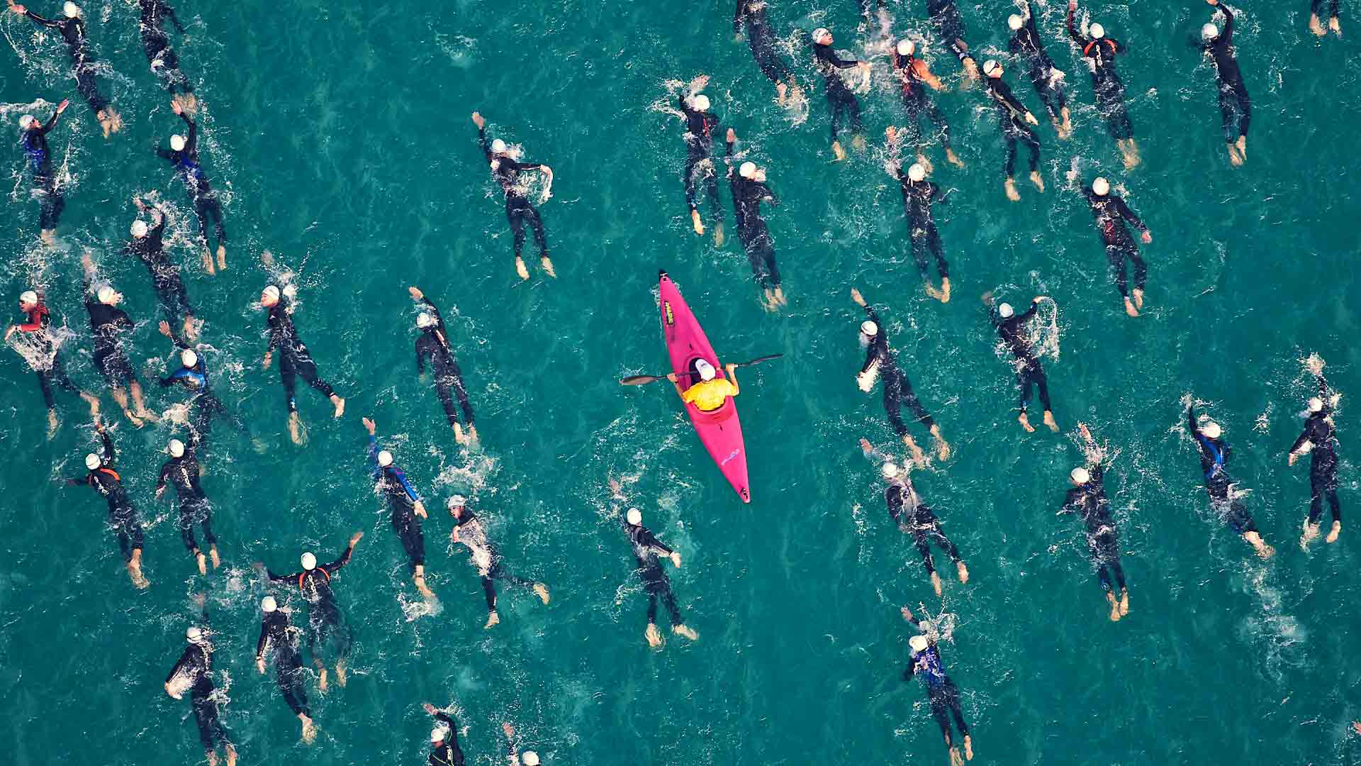 Red canoe pictured from above, surrounded by people swimming in the ocean, intended to symbolise the idea of standign out from the crowd.