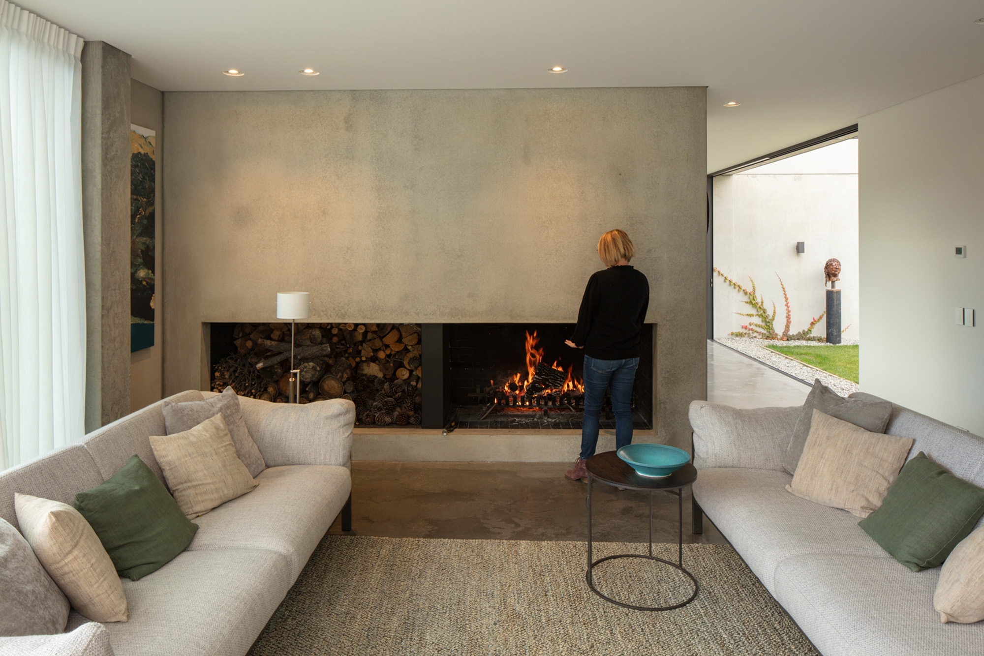 Woman warms her hands at a lounge fire with two white couches and a woven rug.