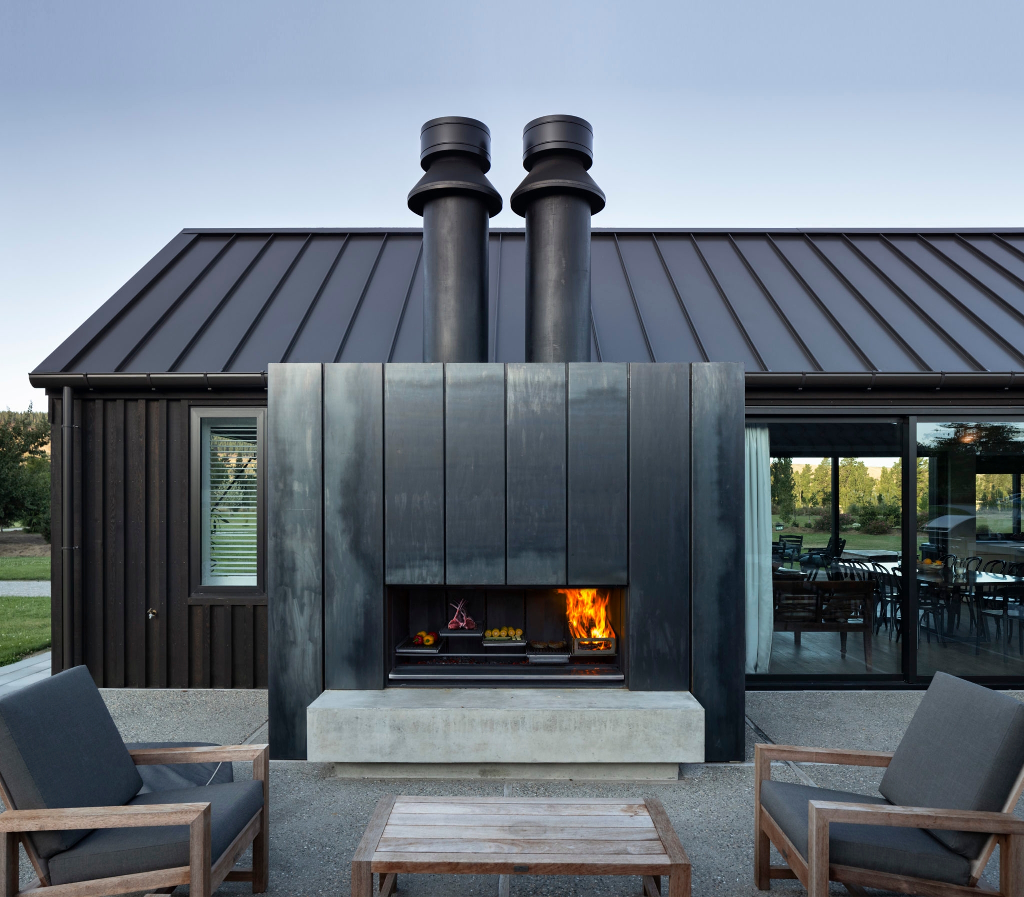 Photo of a modern outdoor patio area featuring a large, industrial-style fireplace with two prominent black chimney stacks extending from the top. The fireplace has a sleek, metal facade with a visible fire burning inside. There are wooden outdoor chairs with dark grey cushions and a matching coffee table arranged around the fireplace. The building behind the patio has a black metal roof and dark vertical siding, with large sliding glass doors leading to an indoor dining area. The overall setting is minimalist and contemporary, with a view of greenery in the background.