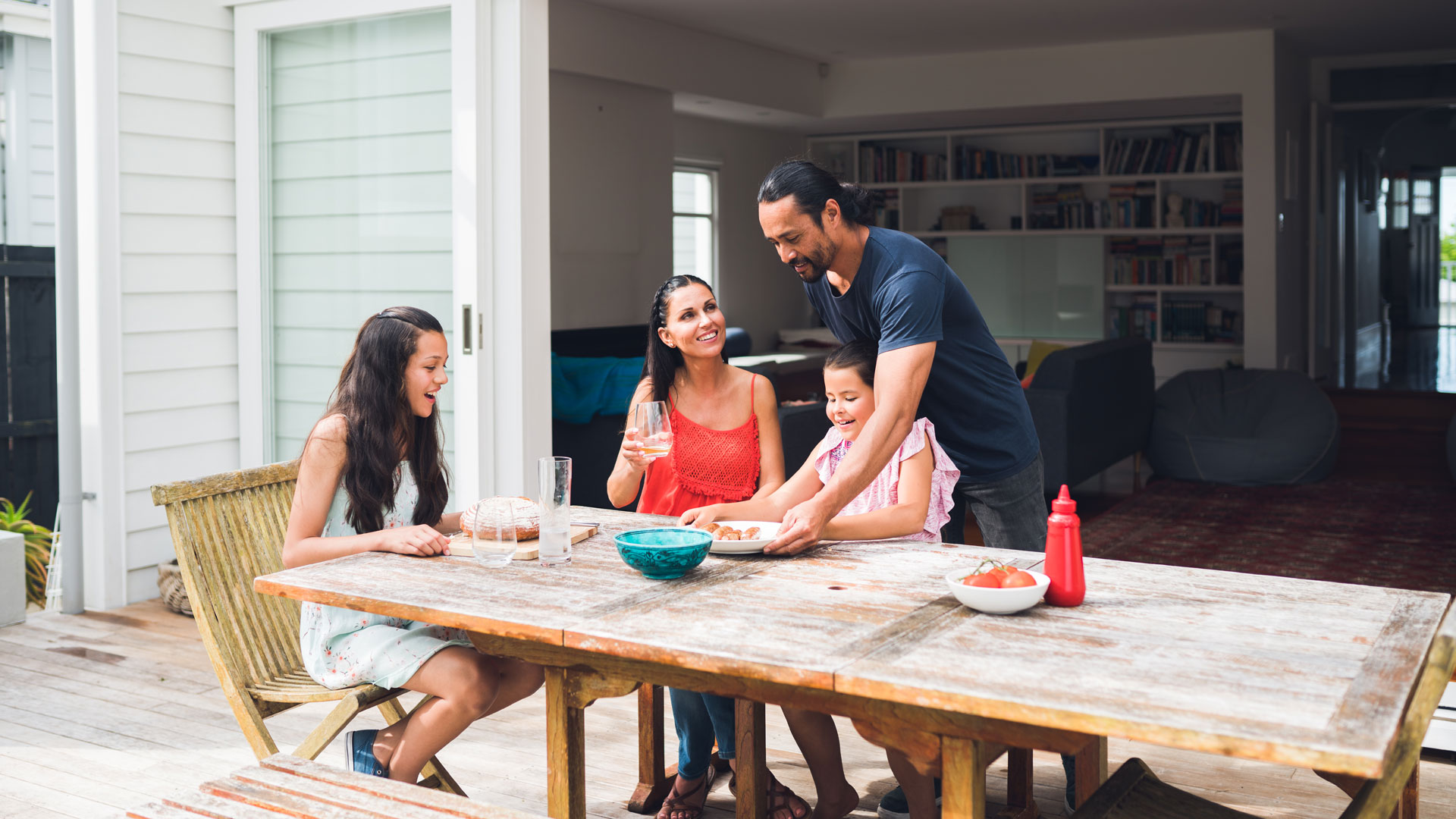 A Kiwi family eating a meal at an outdoor table at their house on the back deck.