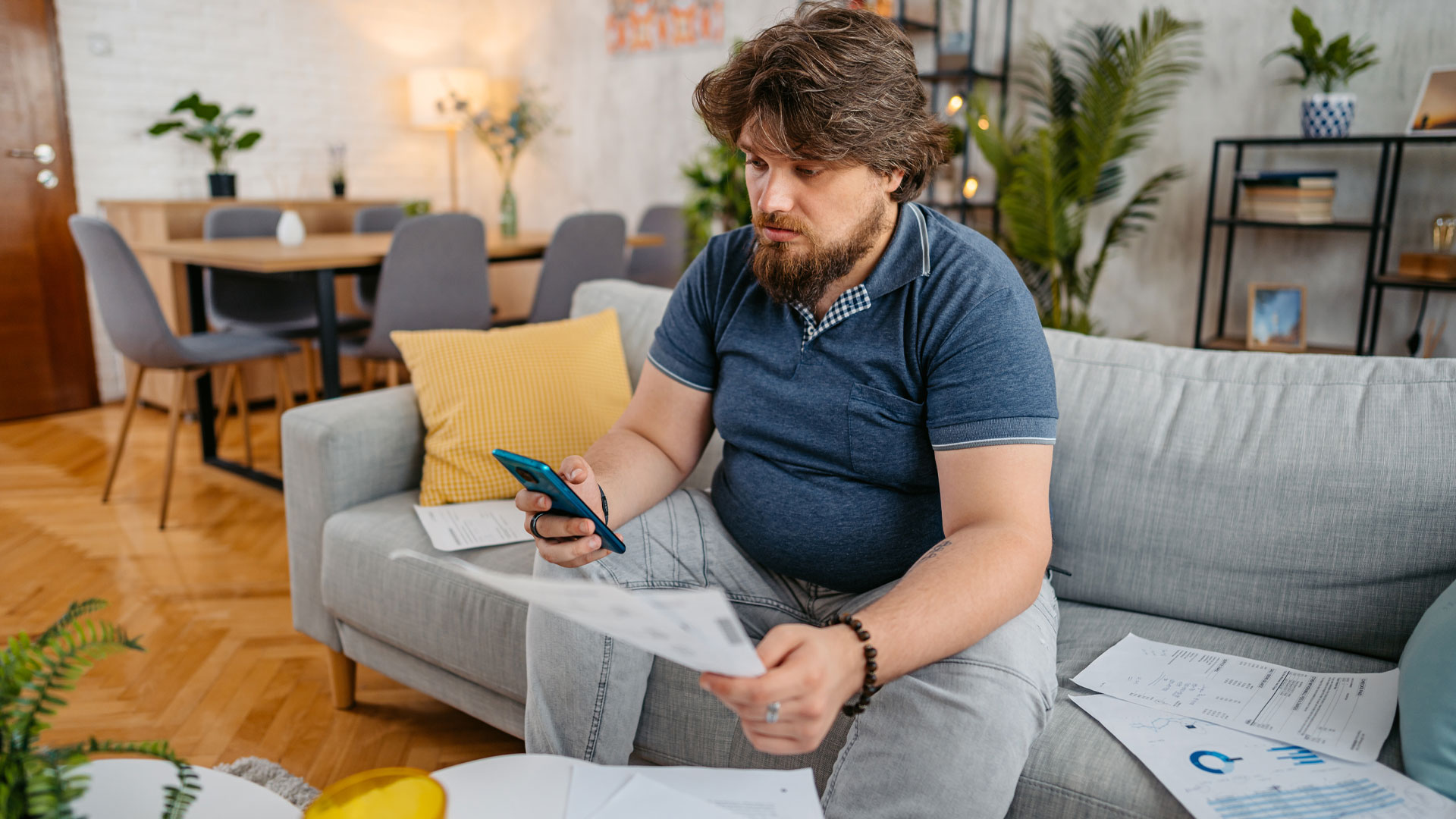 Man sitting on a sofa at home with papers scattered around him and looking at his phone.